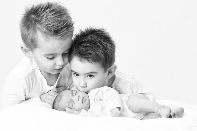 Portrait en noir et blanc en studio de deux enfants et d'un bébé couché sur un drap blanc pour Lorie Diaz photographe