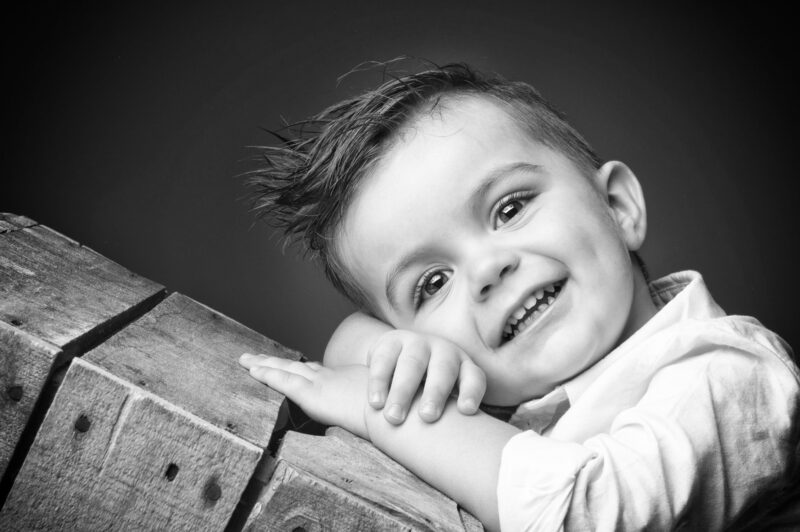 Portrait en studio en noir et blanc sur fond noir d'un petit garçon de face sur une caisse en bois pour Lorie Diaz