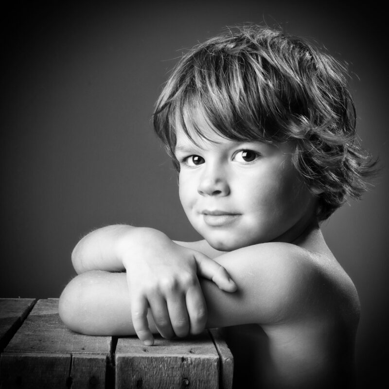 Portrait en studio en noir et blanc sur fond noir d'un petit garçon torse nu appuyé sur une caisse en bois pour Lorie Diaz