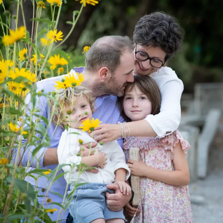 Famille de quatre enlacée en extérieur au milieu de fleurs jaunes : le père tient son jeune enfant, la mère et la grande sœur se serrent contre eux.
